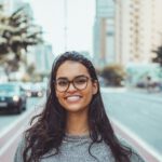 A young woman with glasses smiling on a city street, embracing urban lifestyle.