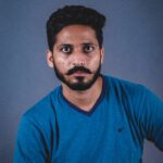 A man with facial hair in a blue shirt poses for a portrait against a gray background.